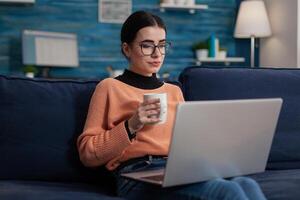Student sitting on sofa holding cup and laptop. Freelance programmer on couch with glasses working from home in living room. Content creator browsing on social media using digital device. photo
