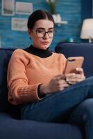 College girl with glasses on sofa looking at smartphone screen. Student on couch reading social media content on mobile phone. Digital influencer checking email on tochscreen device in living room. photo