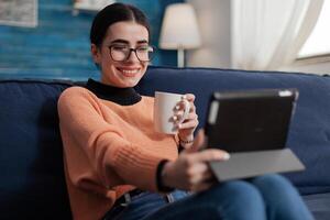 Smiling student on sofa casually talking in videocall using tablet. Content creator with glasses on couch watching on social media. Digital influencer relaxing with cup of coffee or tea. photo