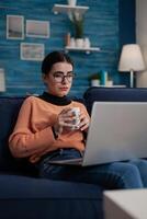 Content creator on couch with laptop on lap holding cup. Student with glasses sitting on sofa learning remote from home in living room. Influencer relaxing looking at screen. photo