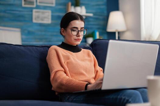 Student influencer on couch holding laptop and typing. Programmer with glasses on sofa writting code in home living room office. Content creator looking at screen posting on social media. photo