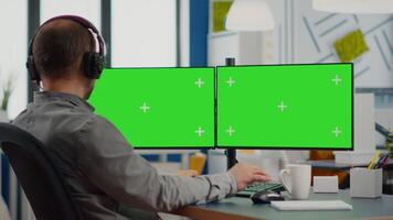 Employee with headphones using dual monitror setup with green screen, chroma key mock up isolated display sitting in production studio. Man ditor processing film montage on pc in creative agency video