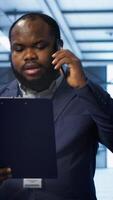 Vertical IT professional looking over data center checkup checklist paperwork, talking on phone in server farm. African american man surrounded by server rigs, chatting during telephone call, camera A video