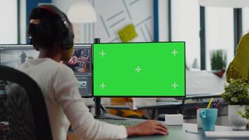 Black woman videographer employee with headphones using computer with green screen, chroma key mockup isolated display sitting in production studio. African editor processing film montage video