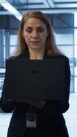 Vertical Software developer walking in data center, overseeing server operations using laptop. Woman ensuring optimal performance and data integrity in server farm using notebook, camera A video