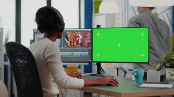 African woman videographer with headset processing film montage working at greenscreen, chroma key isolated display of computer. editor editing movie in post production software in agency office video