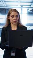 Vertical Software developer walking in data center, overseeing server operations using laptop. Woman ensuring optimal performance and data integrity in server farm using notebook, camera B video
