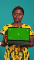 Vertical Young adult showing a laptop with isolated mockup screen in studio, presenting a display with green screen against blue background. Female model posing with chroma key. Camera B. video