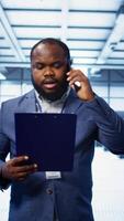 Vertical IT professional looking over data center checkup checklist paperwork, talking on phone in server farm. African american man surrounded by server rigs, chatting during telephone call, camera B video