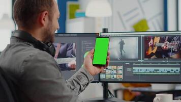 editor falando em ligar segurando Smartphone com verde tela, croma chave zombar acima isolado exibição levando pausa a partir de retocada filme sentado dentro começar acima escritório. equipe trabalhando dentro fundo video