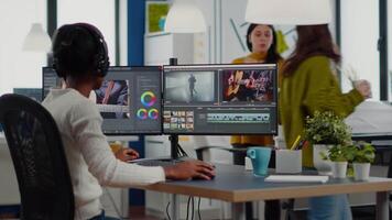 Black videographer putting on headset editing movie using post production software working in creative start up agency office. African editor processing footage on computer with dual displays video