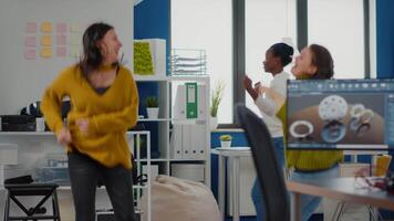 contento diverso donne d'affari danza nel azienda ufficio festeggiare successo di progetto, cantando e avendo divertimento durante Lavorando ore. eccitato multietnico ingegneri utilizzando computer mostrando mascalzone Software video