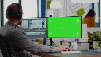 Man videographer with headset editing movie in post production software working at greenscreen, chroma key isolated display of computer. editor processing audio film montage in start up office video