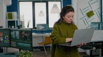 Employee looking at camera smiling standing in front of camera working in creative agency office holding laptop. Videographer works in multimedia studio production editing in modern workplace. video