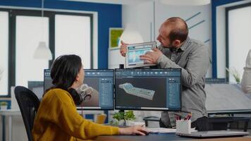 Team of architects and engineers working on building prototype project, using computers running 3D CAD Software. Project manager discussing with employee holding tablet explaining technical specifics video