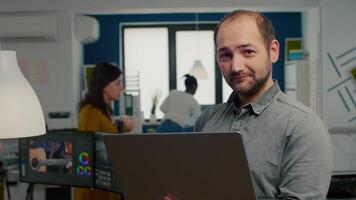 editor worker standing in front of camera smiling working in creative agency office holding laptop. Man videographer works in multimedia studio production editing in modern workplace. video