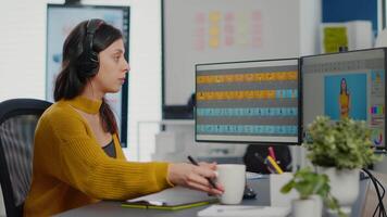 Photo editor with headphones retouching image using stylus pen, drinking coffee, working on PC with two displays. Woman retoucher editing in software app with graphic tablet sitting in creative agency video