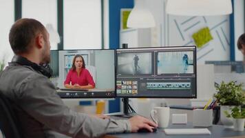 Videographer in web online conference with project manager on call editing client work, getting feedback on commercial movie using post production software on two monitors in start up office video