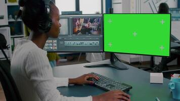 African woman videographer with headset processing film montage working at greenscreen, chroma key isolated display of computer. editor editing movie in post production software in agency office video