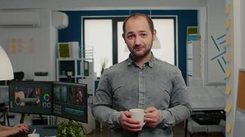 Portrait of videographer standing in start up creative agency office holding cup of coffee looking at camera and smiling. Man working in multimedia studio production editing in modern workplace. video