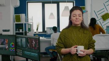 Portrait of creative designer smiling at camera holding a cup of coffee standing in start up agency office. Woman working in multimedia studio production editing in workplace with diverse team. video