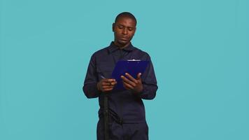 Portrait of security guard holding clipboard, taking notes while overseeing premises. Private law enforcement worker using pen to write down orders received, studio backdrop, camera B video