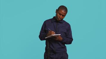 Portrait of security guard using notepad to take notes of suspicious activity while overseeing premises. Private law enforcement worker documenting security protocols, studio backdrop, camera B video