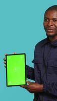 Vertical Portrait of smiling security guard at work showcasing isolated screen tablet while overseeing premises. Joyful private law enforcement worker presenting chroma key device, studio backdrop, camera A video