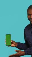 Vertical Portrait of upbeat security staff member showing green screen smartphone. Cheerful patrolling agent maintains secure environment, showcasing mockup phone, studio background, camera A video