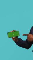 Vertical Joyful security guard working protection job presenting chroma key cellphone. Portrait of merry bodyguard showcasing isolated screen mobile phone, isolated over studio background, camera A video