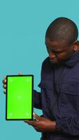 Vertical Happy security guard working protection job presenting mockup tablet. Portrait of cheerful bodyguard showcasing green screen device, isolated over studio background, camera A video