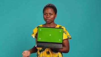 Young adult showing a laptop with isolated mockup screen in studio, presenting a display with green screen against blue background. Female model posing with chroma key. Camera A. video