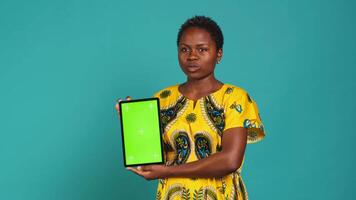 Feminine person in native clothing holds a tablet with mockup screen, creating an advertisement against blue background in studio. Young woman smiling and presenting copy space. Camera A. video