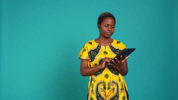 Natural cute girl browsing internet on a tablet against blue background, navigating on online web pages by using a portable device. Woman posing with a gadget and traditional outfit. Camera B. video