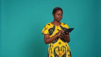 Happy positive adult navigating on online webpages in studio, using portable gadget to browse things on internet connection. Woman in traditional ethnic attire poses against background. Camera B. video