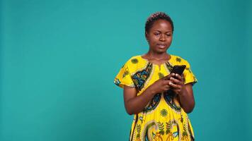 Native girl in ethnic outfit browsing internet webpages in studio, using her smartphone to check texts and social media platforms. Young person standing against blue background. Camera B. video