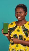 Vertical Young confident woman presenting a green screen on smartphone, working in studio on an advertisement. Smiling natural girl holds a phone with isolated chroma key display. Camera A. video