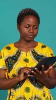 Vertical Natural cute girl browsing internet on a tablet against blue background, navigating on online web pages by using a portable device. Woman posing with a gadget and traditional outfit. Camera A. video