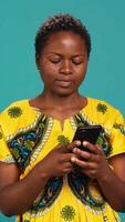Vertical Native girl in ethnic outfit browsing internet webpages in studio, using her smartphone to check texts and social media platforms. Young person standing against blue background. Camera A. video