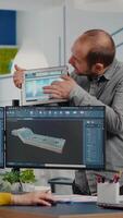 Team of architects and engineers working on building prototype project, using computers running 3D CAD Software. Project manager discussing with employee holding tablet explaining technical specifics video