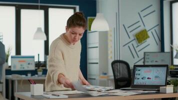 Busy woman in startup office holding clipboard looking at papers with sales charts at desk. Business analyst working hard searching for turnover data statistics with laptop showing profit analytics. video