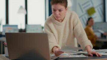Focus on laptop in front of startup employee looking and charts comparing sales statistics standing at desk. Woman in busy office sorting through business papers looking at data analytics. video