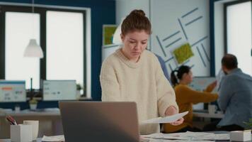 Portrait of focused startup employee looking at laptop and charts comparing sales statistics standing at desk. Woman in busy office sorting through business papers looking at data analytics. video