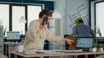 Busy woman in startup office talking on smartphone looking at papers with sales charts at desk. Business analyst using mobile phone searching for data statistics with laptop showing profit. video