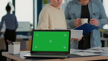 Laptop computer with green screen on desk in front of startup colleagues talking about sales charts papers on clipboard. Focus on portable computer screen with chroma key empty background. video