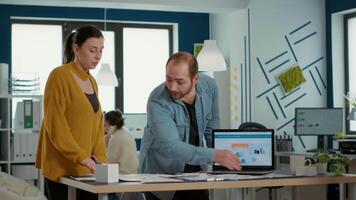 Startup office worker explaining sales startegy to new employee using laptop with business analytics and turnover charts. Man standing at desk talking about marketing with woman doing internship. video