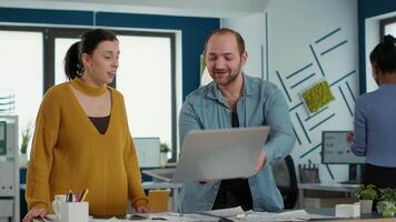 leende medarbetare i kontor ser på bärbar dator skärm jämförande företag analys med försäljning diagram stående på skrivbord. kollegor håller på med lagarbete arbetssätt på framgångsrik projekt använder sig av bärbar dator. video