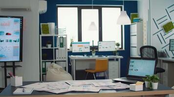 Empty startup office with laptop showing analytics on display with moving charts and tv wall screen with statstics. Interior of marketing department desk full of business papers with financial data. video