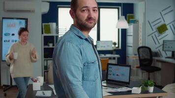 Portrait of confident man entering frame posing with arms crossed and smiling in busy startup office. Relaxed employee acting casual looking at camera in marketing department of small company. video