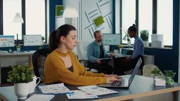 Focused startup employee typing using laptop keyboard while looking at screen and smiling sitting at desk in busy office. Casual business woman working relaxed with papers with charts and statistics. video